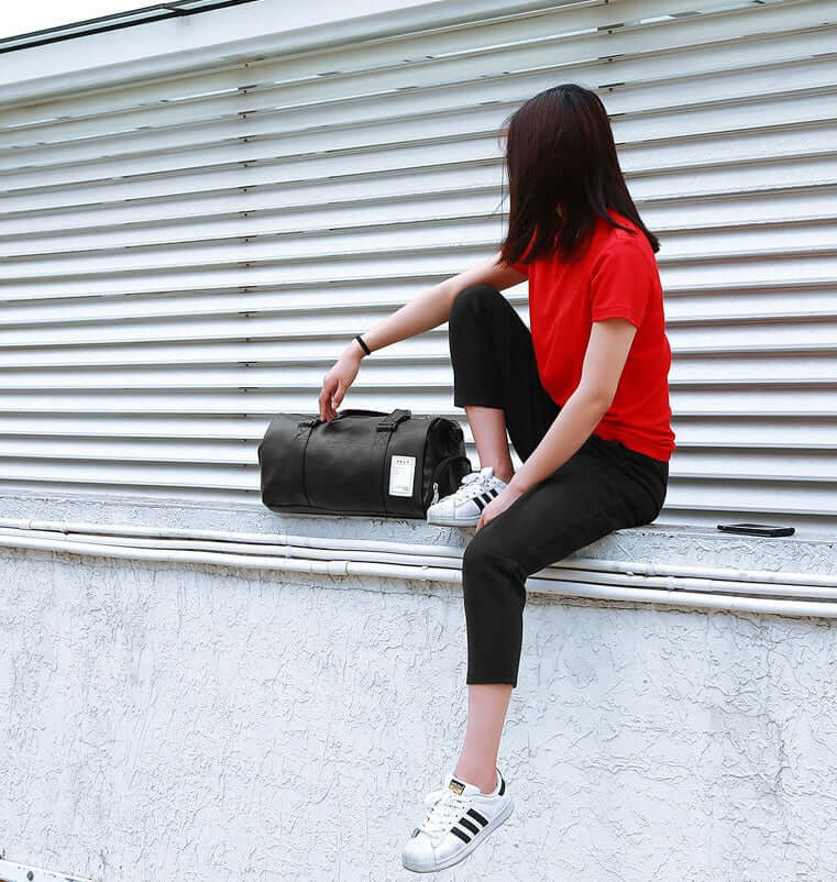 Person in a red shirt sitting on a ledge with a black sports fitness bag, wearing casual sneakers.